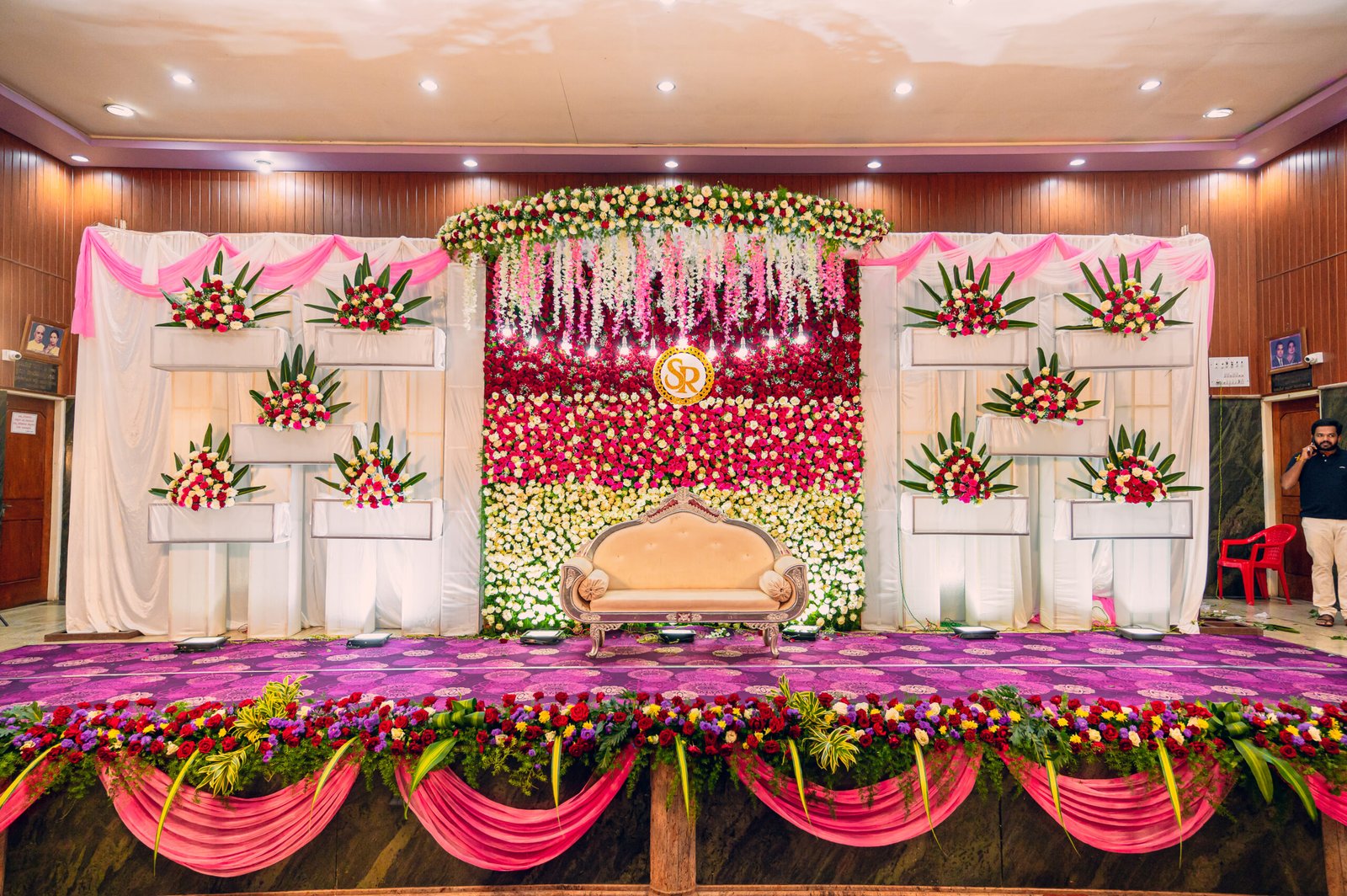 Wedding reception and Mandap stage decorated with a full flower wall backdrop, hanging wisteria, sofa seating, and colourful floral arrangements inside a banquet hall